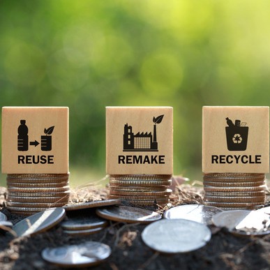 Auf drei Münzstapeln steht jeweils ein Holzwürfel mit der Beschriftung „Reuse“, „Remake“, „Recycle“.