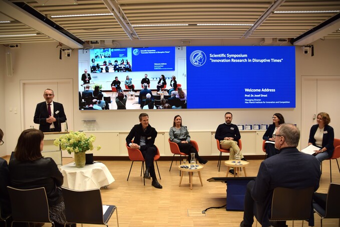 Podiumsdiskussion bei einem Eröffnungssymposium mit mehreren sitzenden Personen und einer stehenden Person, die spricht, vor zwei großen Bildschirmen mit Präsentationsfolien.