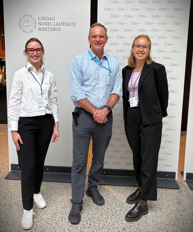 Drei Personen stehen vor einer Wand mit dem Schriftzug 'Lindau Nobel Laureate Meetings' und einem Logo, zwei Frauen und ein Mann, alle mit Namensschildern.