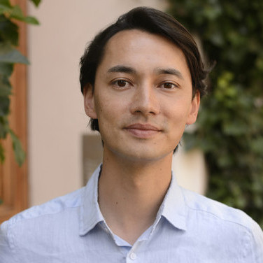 Junger Mann mit dunklem Haar trägt hellblaues Hemd, steht vor einer Wand mit grünen Pflanzen.