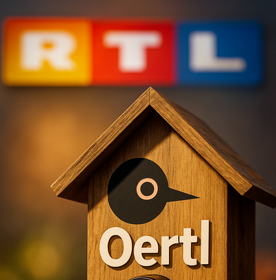 Symbolbild: Holz-Vogelhaus mit Kamera und der Aufschrift ‚Oertl‘ im Vordergrund, im Hintergrund unscharf das Logo des Fernsehsenders RTL.